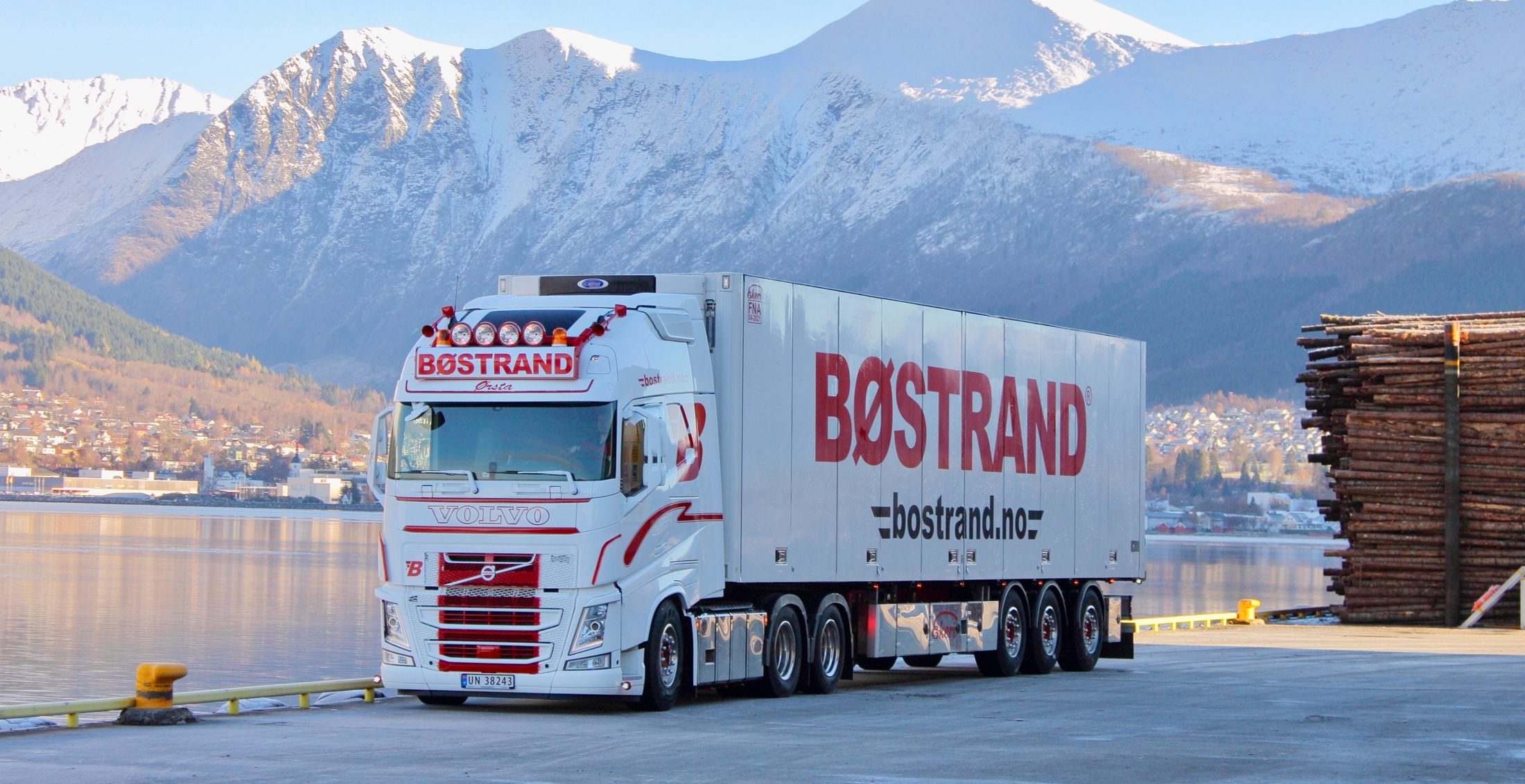 Bøstrand-vogntog parkert ved fjorden med fjell og bygd i bakgrunnen